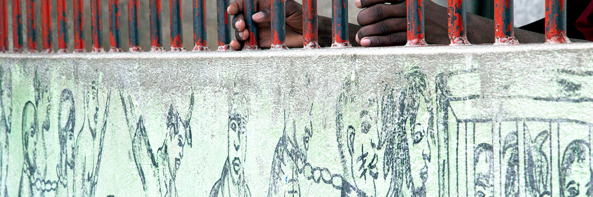 An inmate grasps the bars of an outside wall at the Penitentiary on which a mural of prisoners has been drawn. Port-au-Prince, Haiti. UN Photo/Victoria Hazou