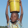 A little boy holds a bucket of potatoes on his head.